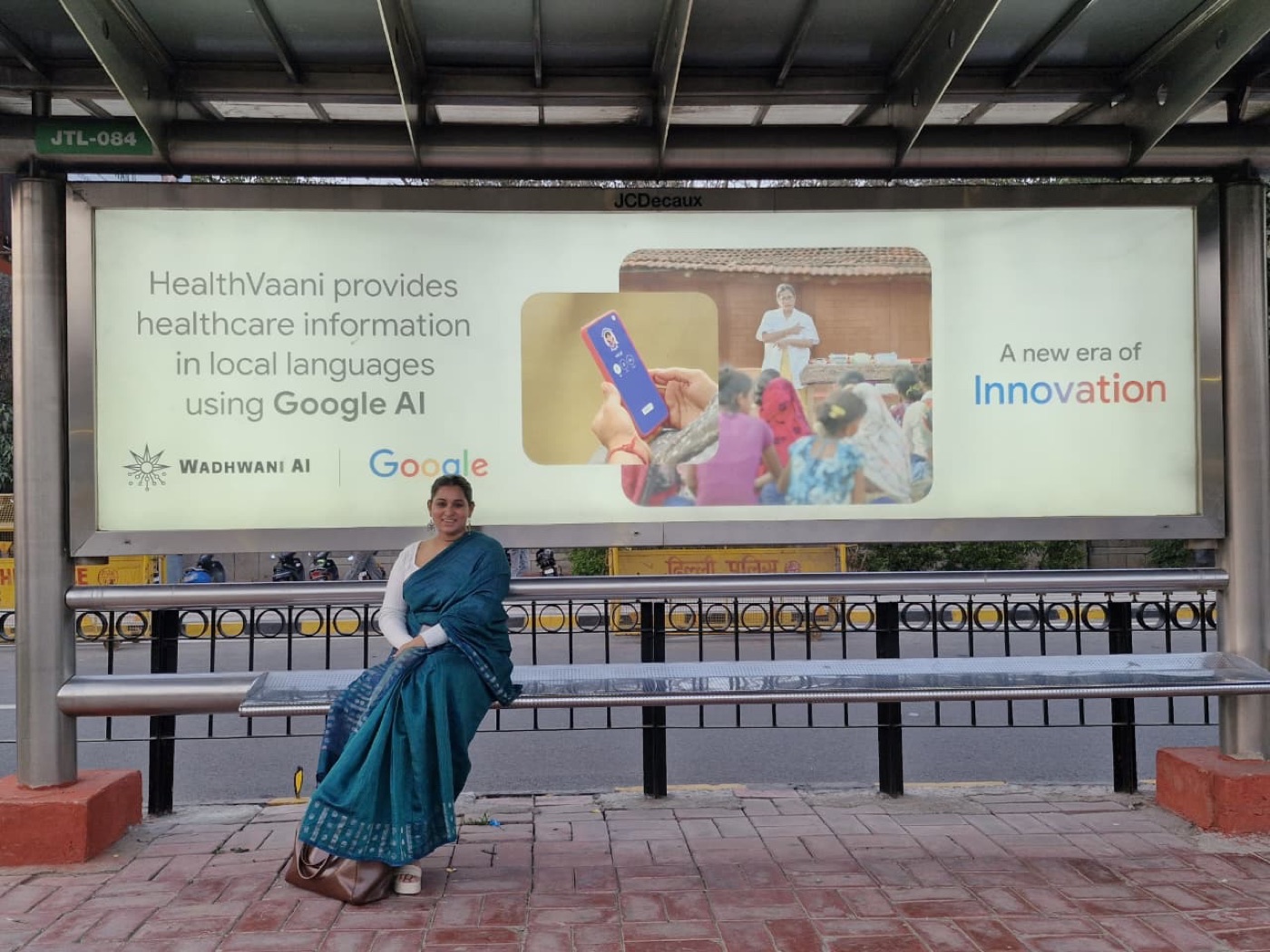 Daksha Dixit standing in front of a Delhi bus-shelter billboard for HealthVaani, co-branded Wadhwani AI and Google, reading "HealthVaani provides healthcare information in local languages using Google AI."